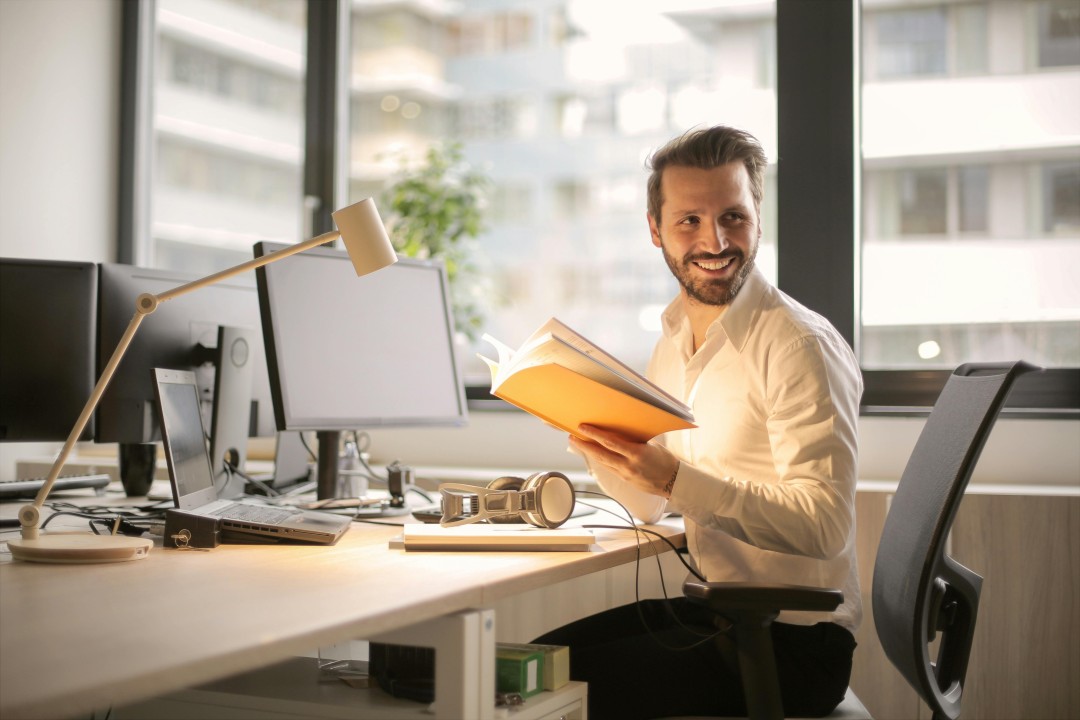 Hombre profesional sonriendo mientras sostiene y revisa un cuaderno en su escritorio de oficina junto a una computadora portátil.