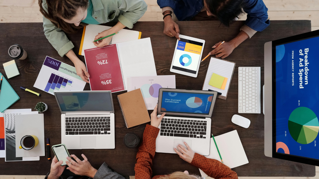Vista superior de un equipo de trabajo colaborando en una mesa con ordenadores portátiles, informes impresos y una pantalla con métricas de presupuesto publicitario.